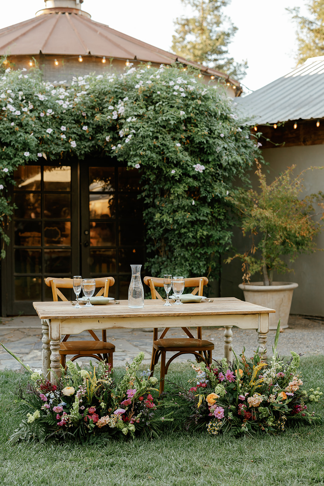 A sweetheart table set outdoors on the grass at Pageo Lavender Farm featuring a summer floral design by Bloom & Forage. The floral styling incorporates a mix of vibrant seasonal blooms arranged at ground level, creating a relaxed and colorful summer reception aesthetic that complements the natural outdoor setting.